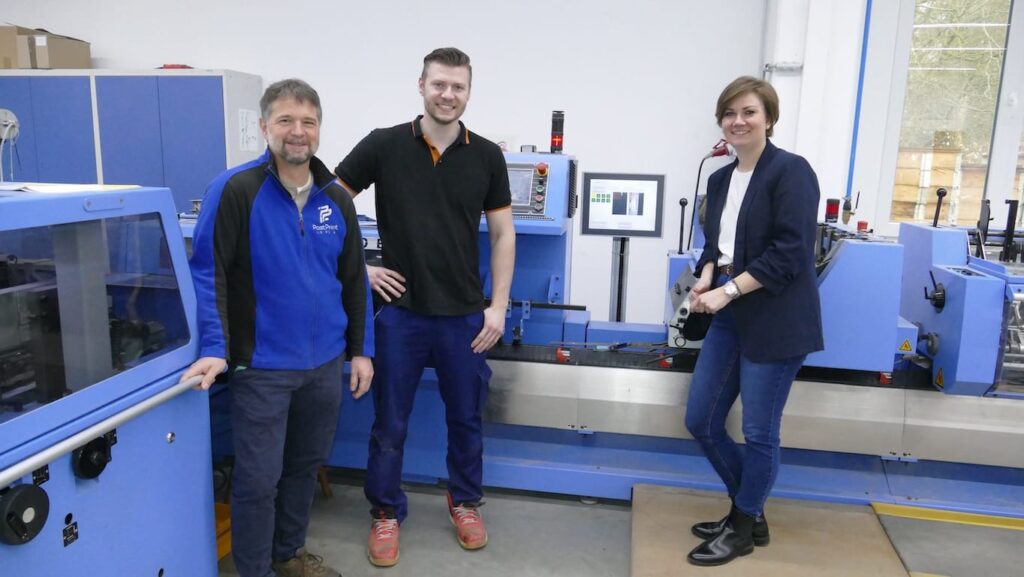 Drei Personen stehen vor einer blauen Weiterverarbeitungsmaschine in der Werkstatt